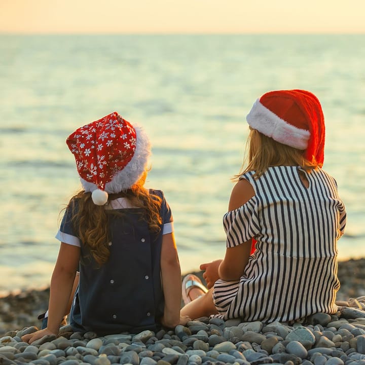 Two young girls sitting my the beach with Santa hats on. 